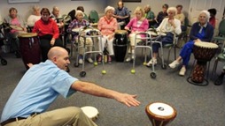 A man reaches for a drum to his right while a audience of nine seniors watch.