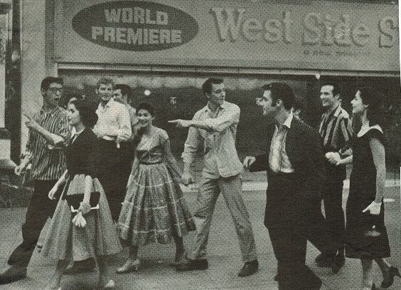 Actors and actresses from West Side Story walk on the sidewalk outside of the theatre.