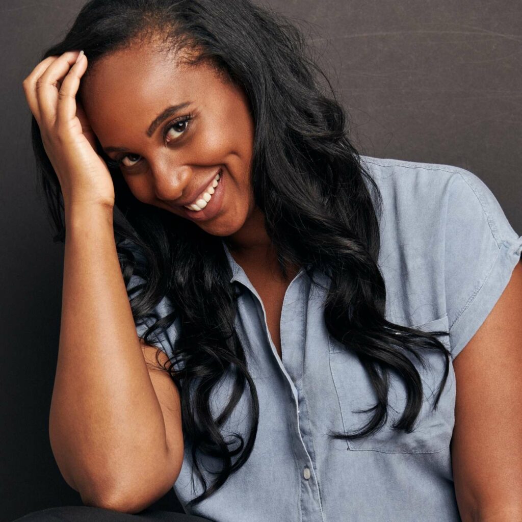 Smiling photo of a Black woman with long black hair wearing a denim button down short sleeved shirt