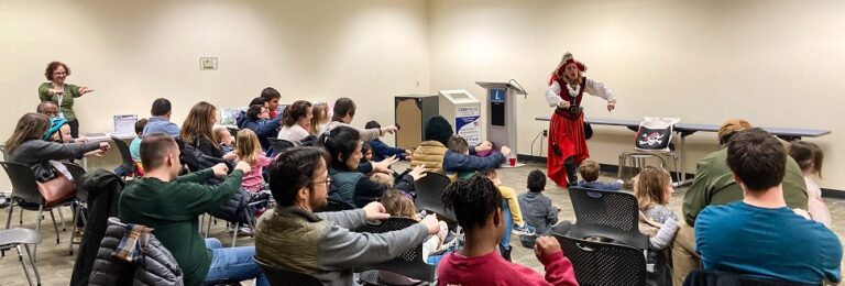 A performer in a red pirate costume leading an audience of small children and adults to mime rowing a boat.