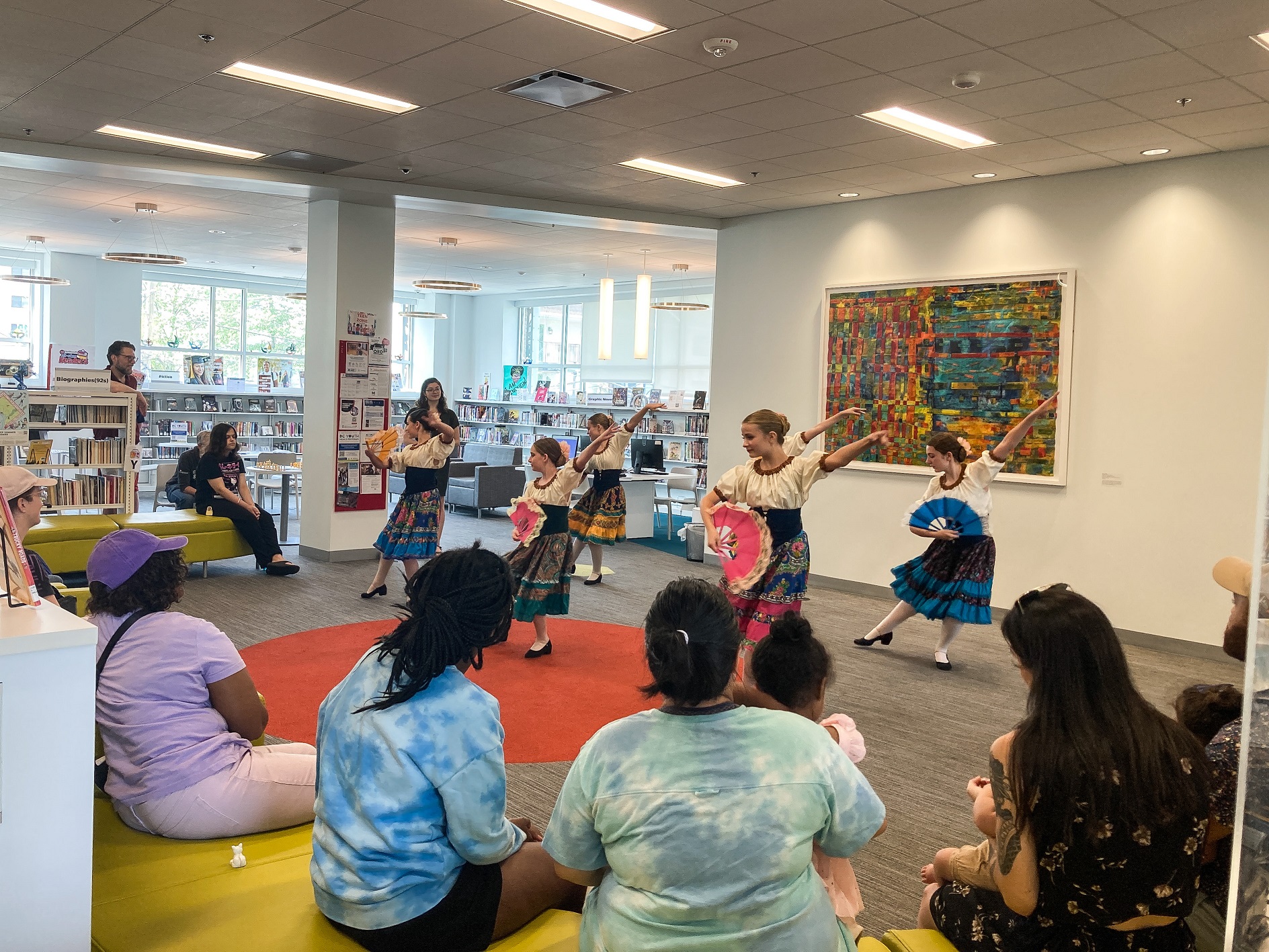 Girls in Spanish dresses dance with fans in a library space as parents and children look on.