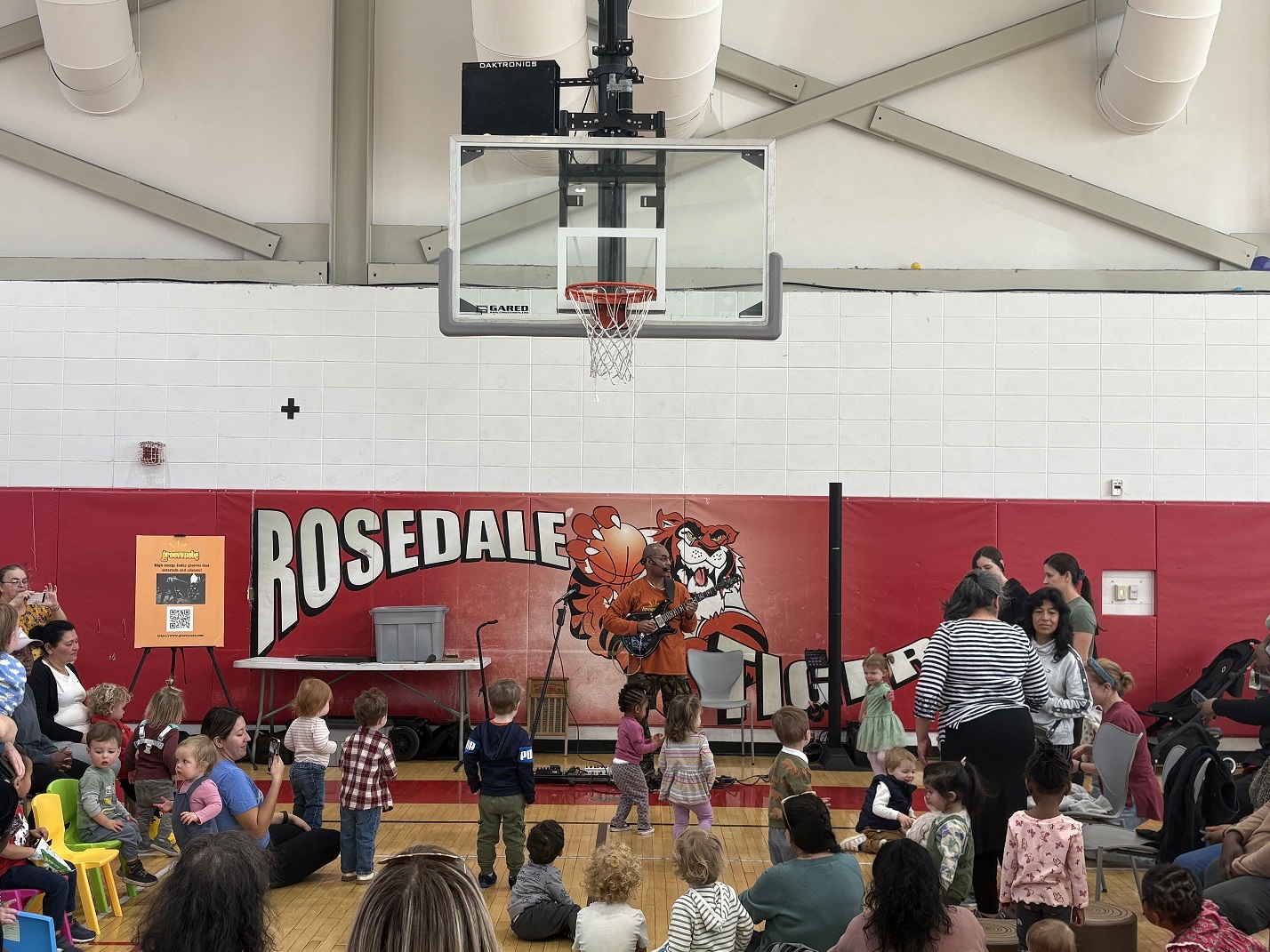 A man with a guitar wearing an orange shirt plays in a basketball court surrounded by children and adults.