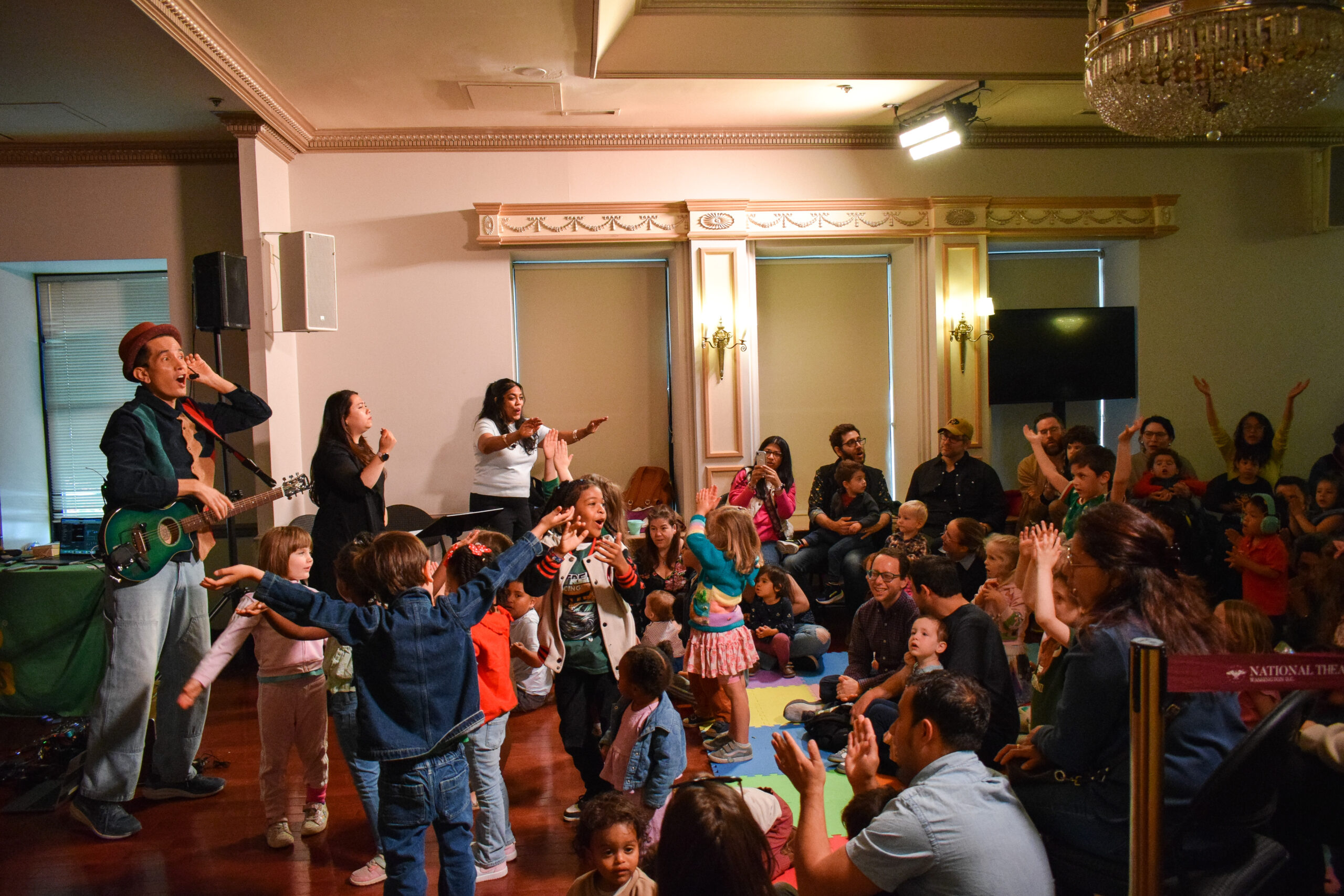 A performer in a fedora and green top gestures for an assembled crowd of children and caregivers to get louder.