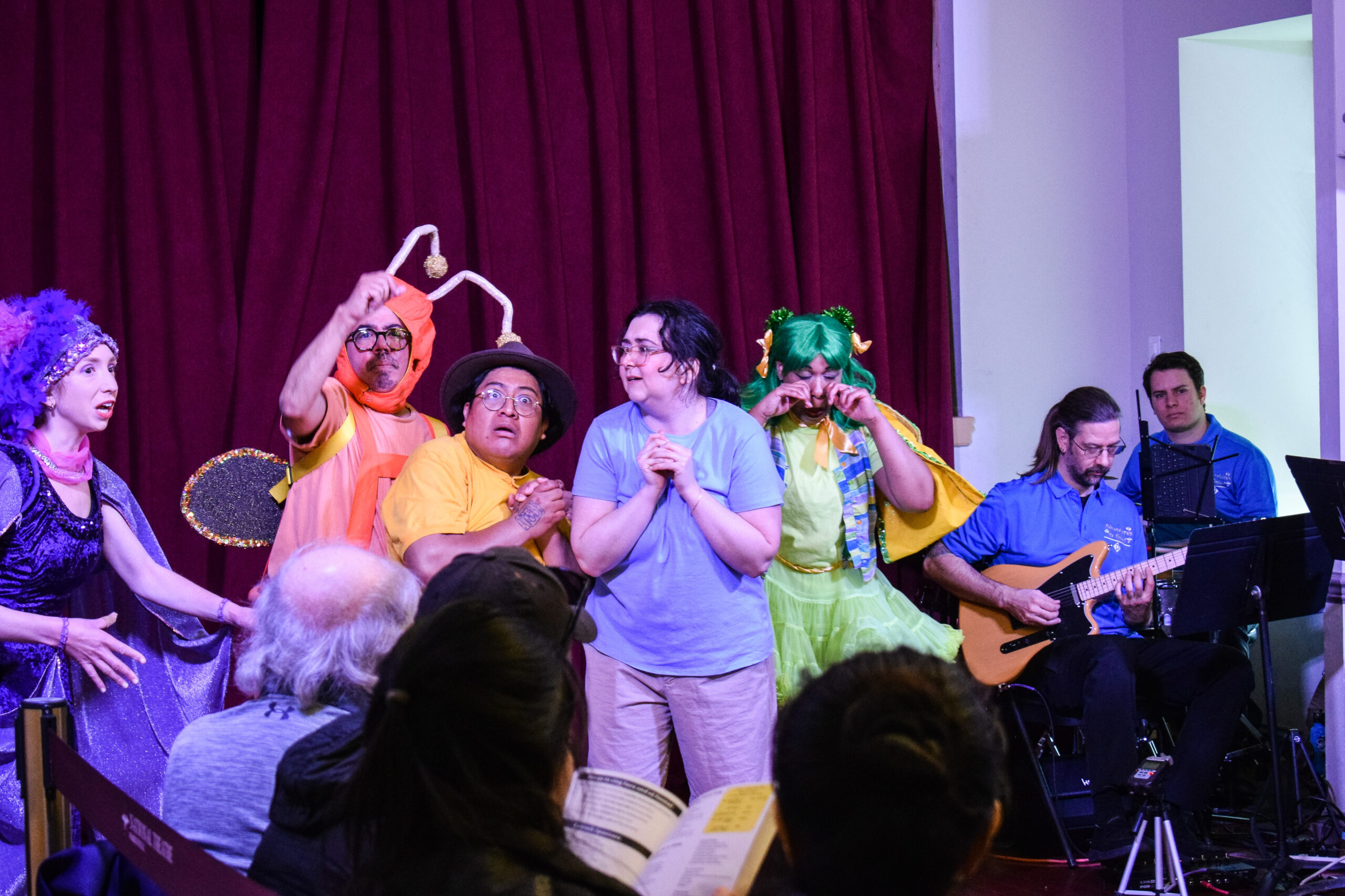 An assortment of characters in bug costumes and contemporary clothes huddles together fearfully in front of a dark red curtain.
