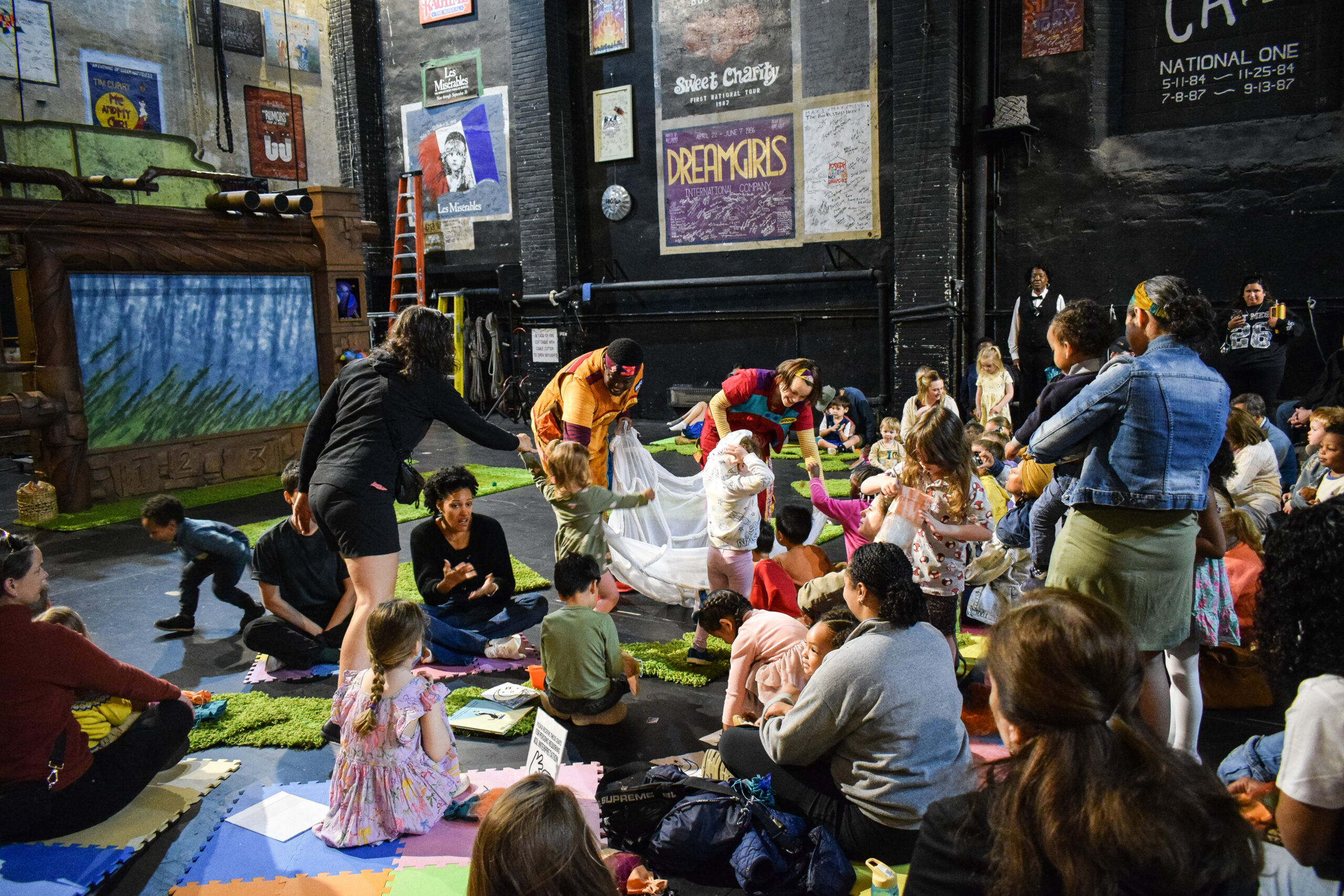 Two performers in orange and red jumpsuits help children gather items into a white net. They are surrounded by posters of previous productions in The National Theatre's history.