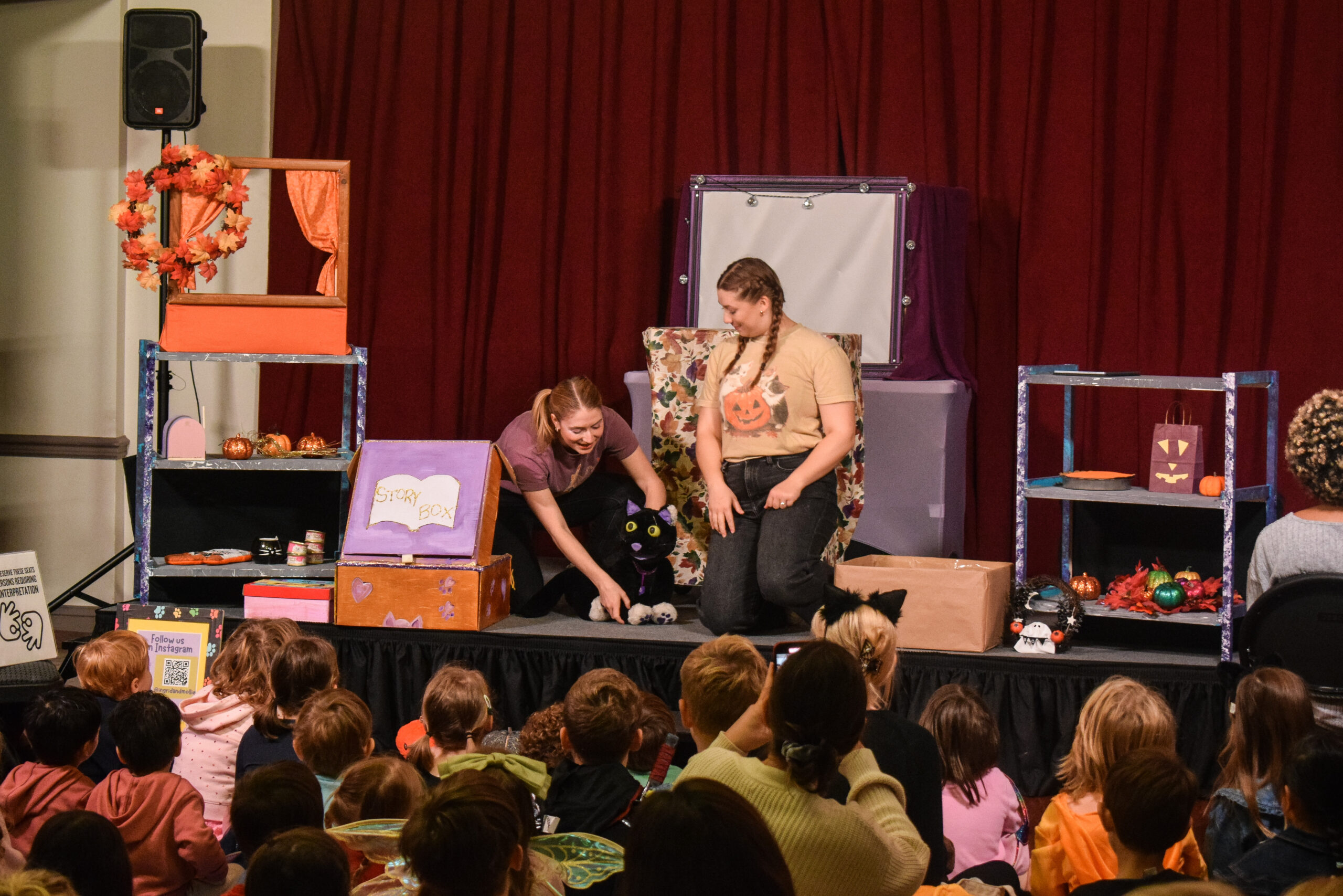 Two women kneel on a stage while operating a black cat puppet. They are surrounded by a set and watched by children seated on multicolored mats.