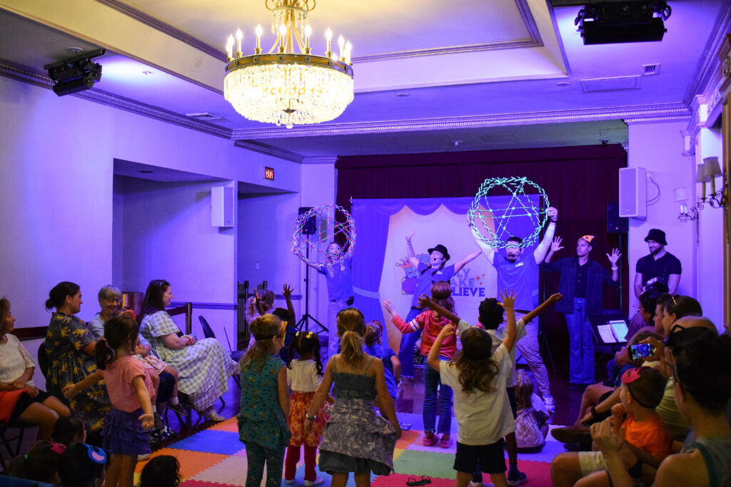 Three performers in blue shirts gesture flamboyantly under blue lighting. Assembled children move along with them while standing on multicolored mats.