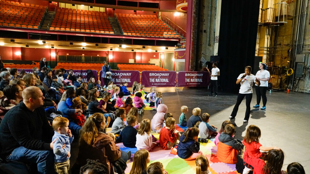 A mass of children and caregivers seated on multicolored mats watch performers in white t-shirts and black pants. The auditorium of The National Theatre is behind them.