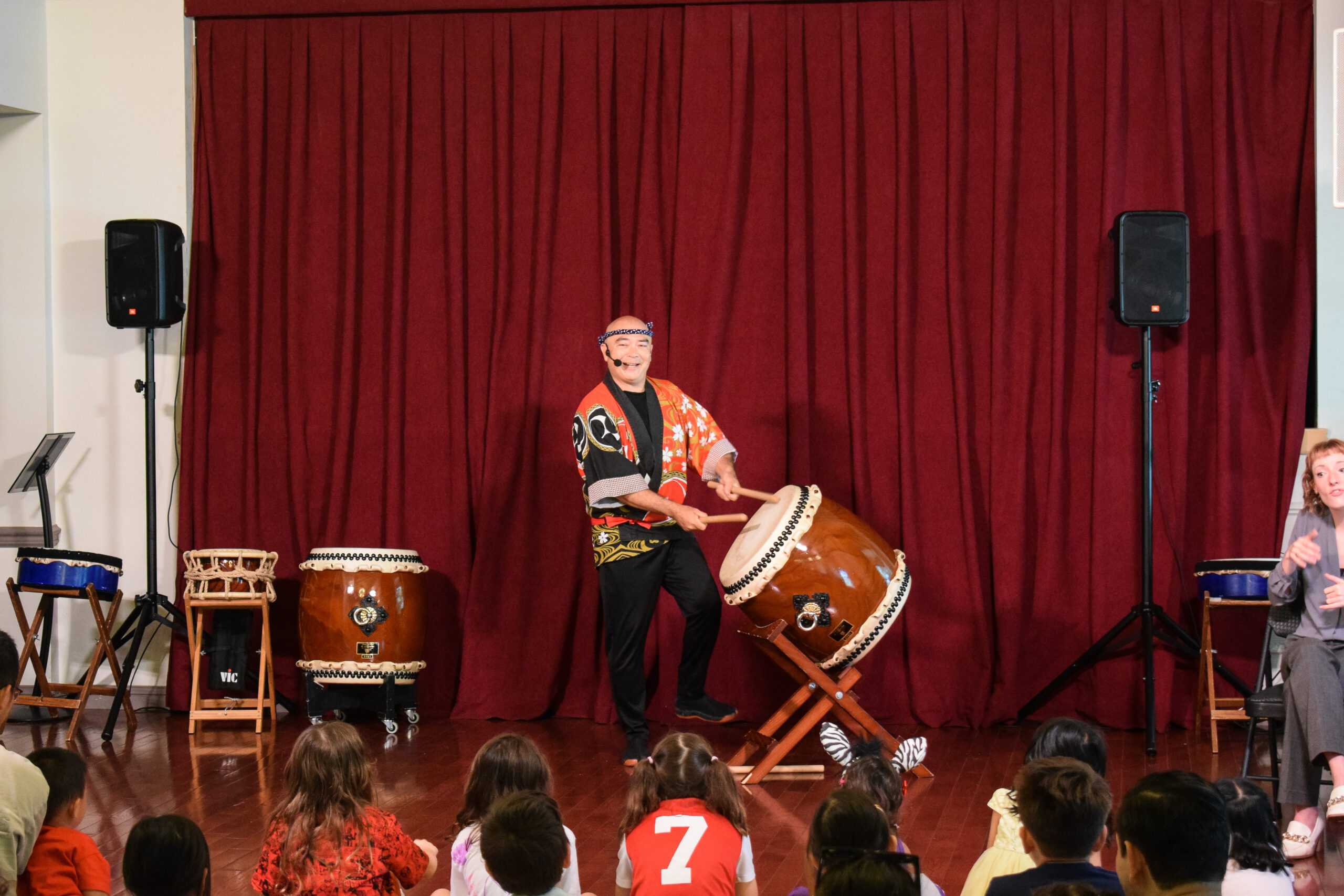 A man in traditional red Japanese garb plays taiko while speaking to an assembled audience of children.