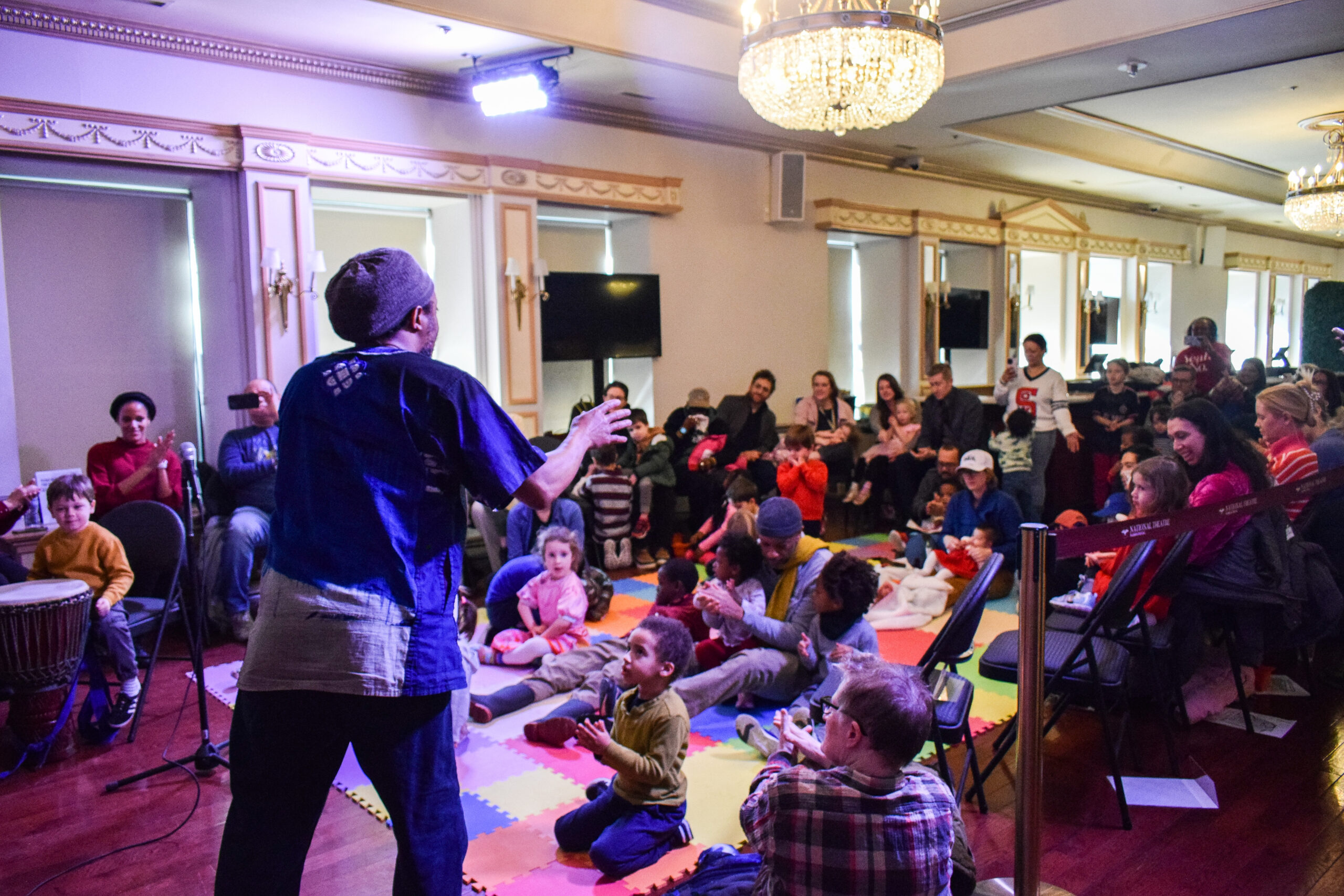 A man in a blue top instructs children and caregivers seated on multicolored mats.
