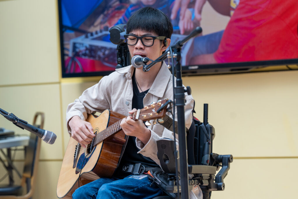 A man in glasses, seated in a wheelchair, sings into a microphone while playing an acoustic guitar.