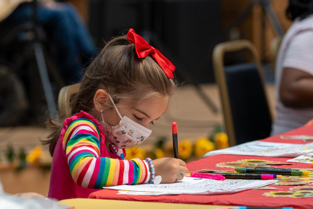 A young girl with a red bow in her hair and a mask on her face colors on a coloring page.