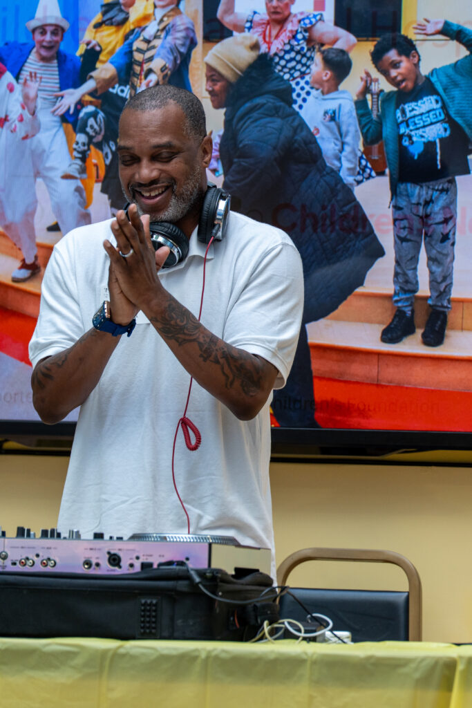 A man in a white t-shirt, wearing a pair of headphones, claps his hands and smiles while standing over his mix station.