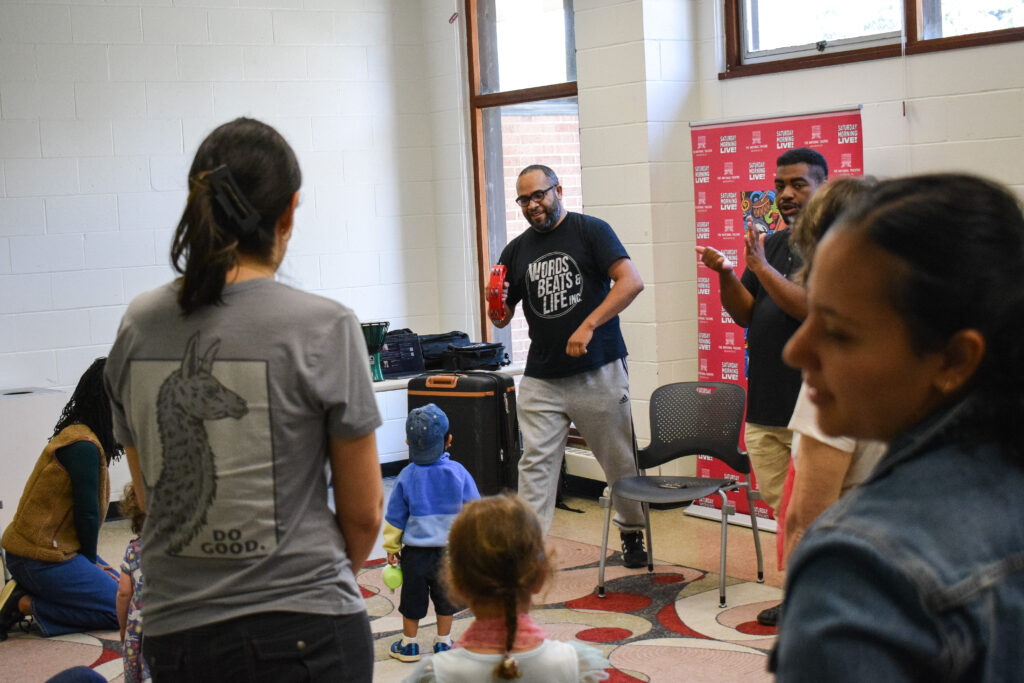 A man in a black shirt instructs children and parents in how to keep a beat.