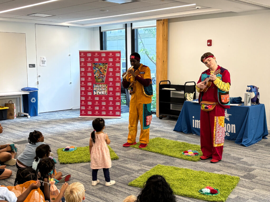 Two men in jumpsuits play instruments in front of a crowd of small children.